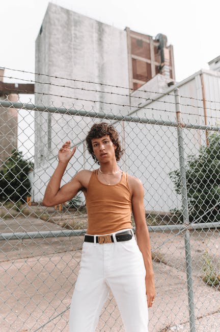 Fashionable adult in a brown top and white pants posing by an urban fence outdoors.