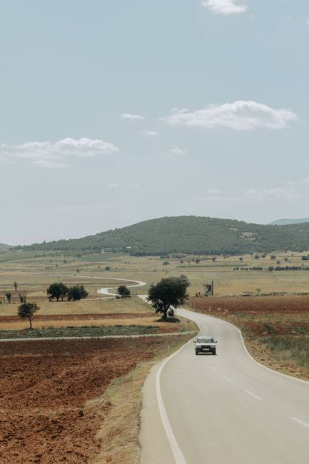 Long winding road through rural landscape with blue sky and mountains in the background.