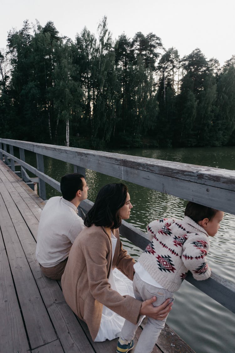 Family With Child Sitting On Bridge Near Water