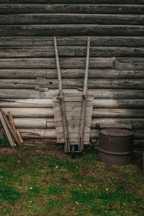 Free An old wooden wheelbarrow leans against a rustic log cabin wall with grass at its base. Stock Photo