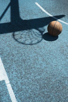 Basketball resting on an urban court next to a hoop shadow in daylight.