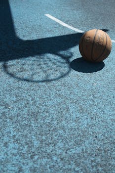 Basketball lying on blue court with hoop shadow, showcasing sports equipment and shadows.