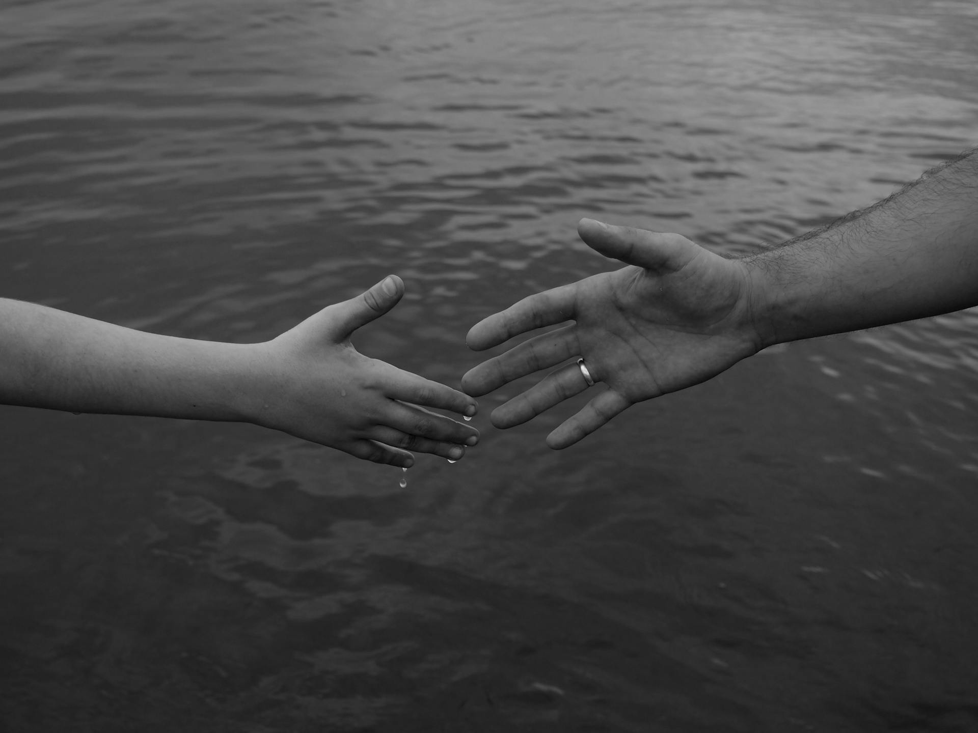 Monochrome image of two hands reaching out over water, symbolizing connection and support.