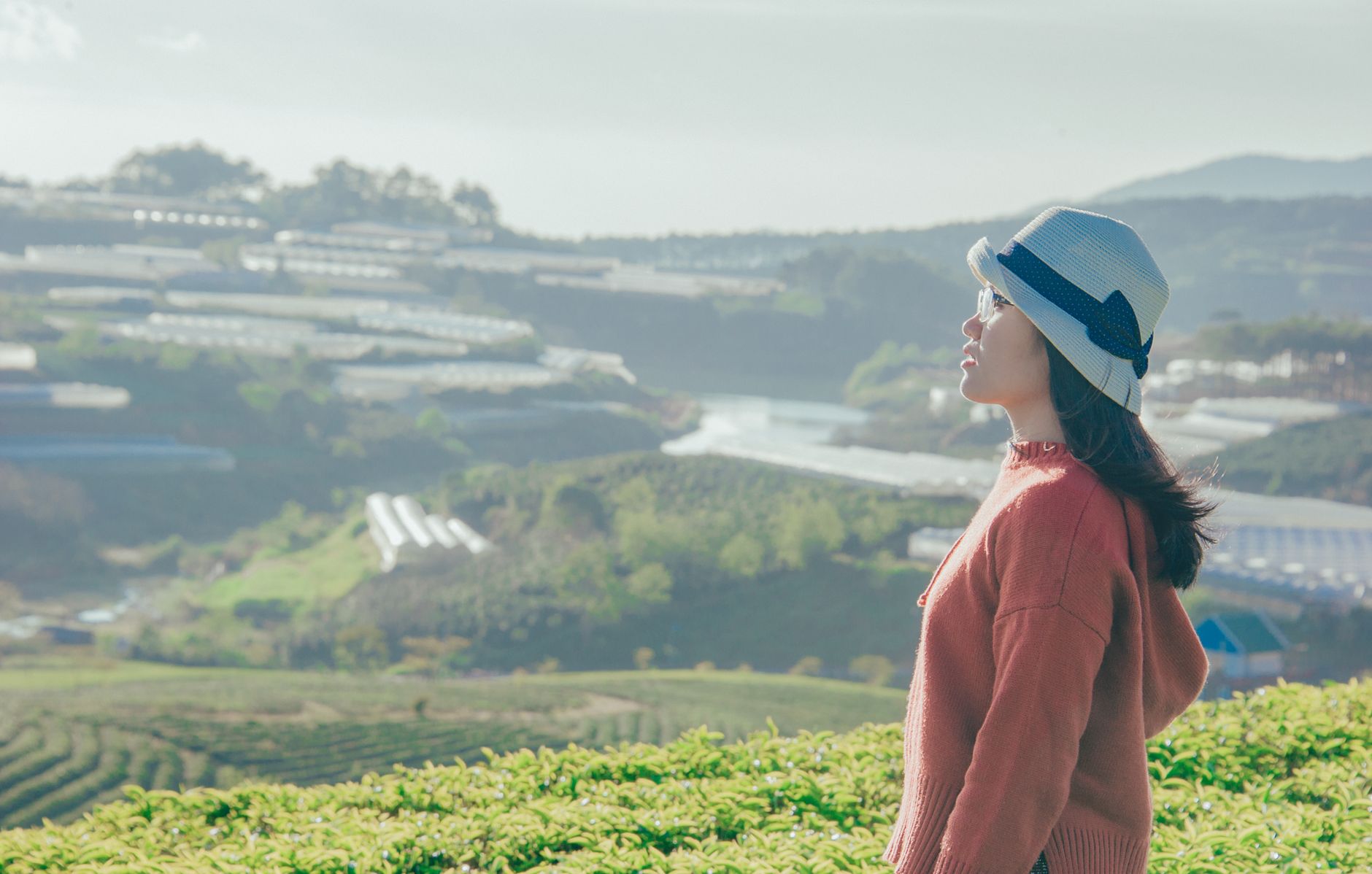 A woman in a hat gazes at an expansive farmland landscape under bright daylight.