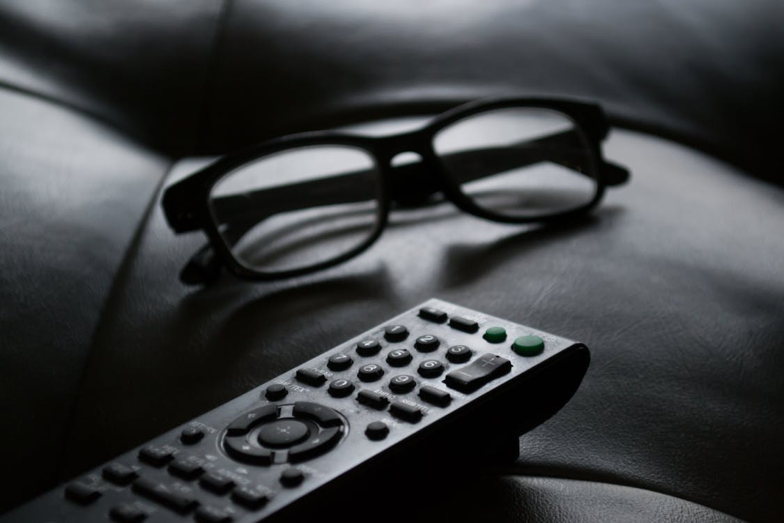Grayscale Photo of Remote Control Near Eyeglasses