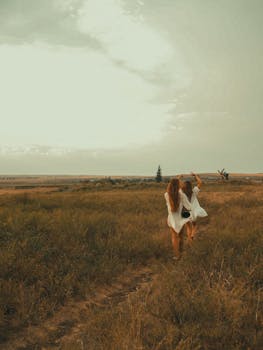 Two women in white dresses walk through a vast grassland under a cloudy sky.