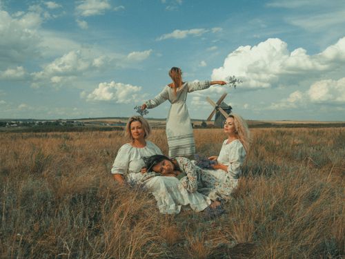 Free Four women in boho dresses relaxing in a tranquil wheat field, with a wooden windmill in the background. Stock Photo