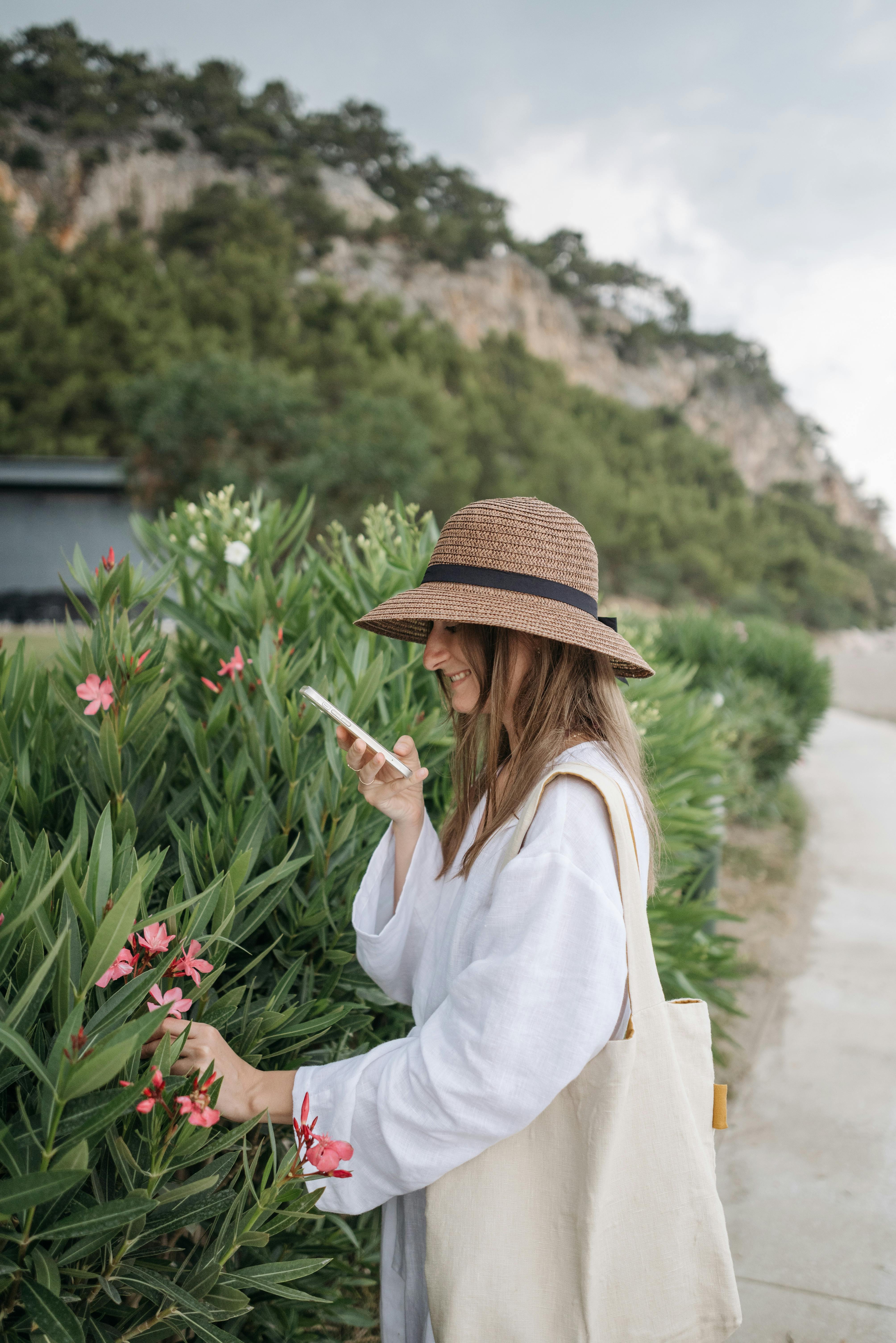 Woman in a straw hat photographing pink flowers with a smartphone in a garden setting.