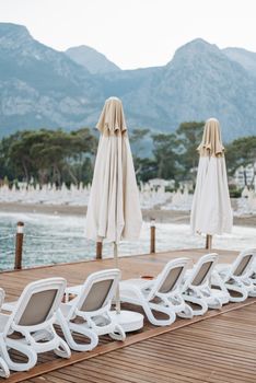 Tranquil resort setting with mountains, empty sunloungers, and closed umbrellas by the beach.