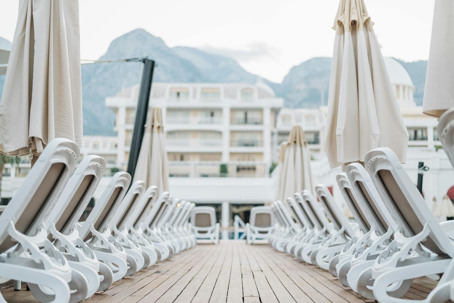 A serene view of a luxury resort's wooden deck lined with sun loungers and umbrellas against a mountain backdrop.