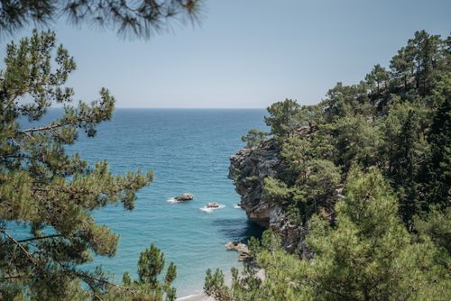Free Stunning view of rugged cliffs and vibrant ocean surrounded by lush green trees on a sunny day. Stock Photo