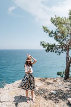 Woman in summer attire taking a selfie on a rocky cliff by the sea, capturing a serene oceanic landscape.
