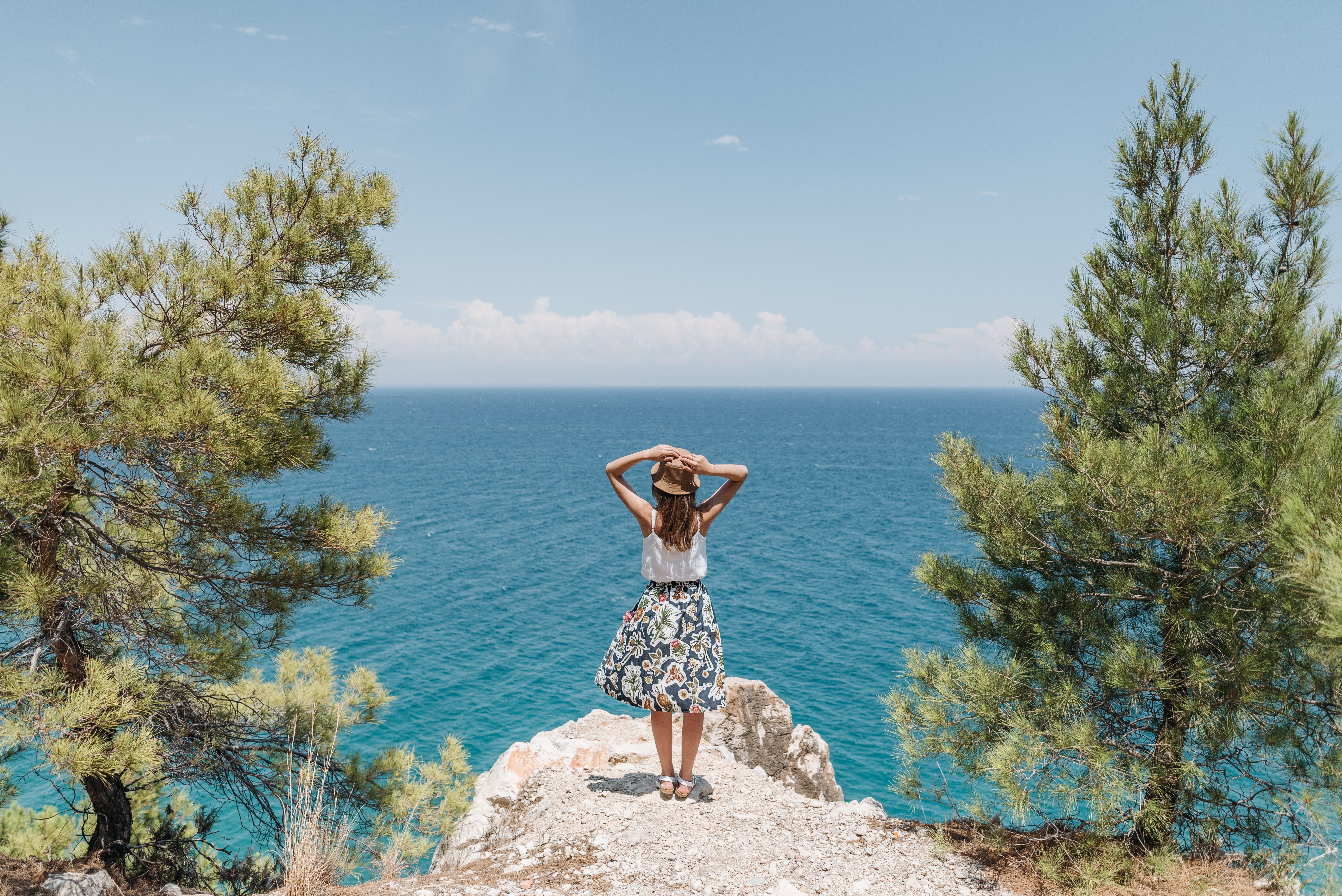 A woman stands on a cliff overlooking the ocean, embracing nature on a sunny summer day.
