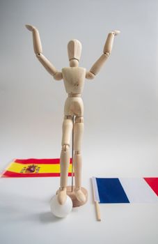 Wooden mannequin with raised arms between Spanish and French flags.