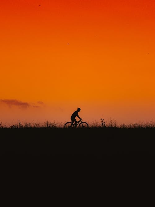 A Silhouette of a Man Riding a Bicycle on Grass Field Under the Sunset Sky