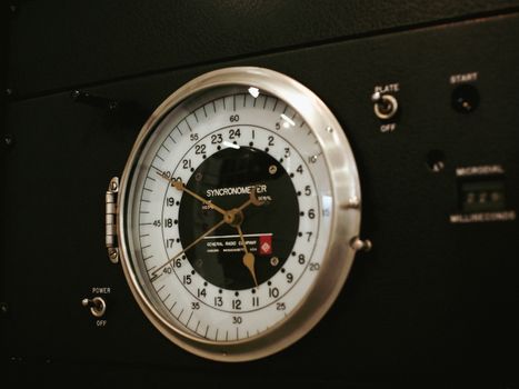 Close-up view of a vintage syncronometer with metal casing and glass cover in an industrial control panel.