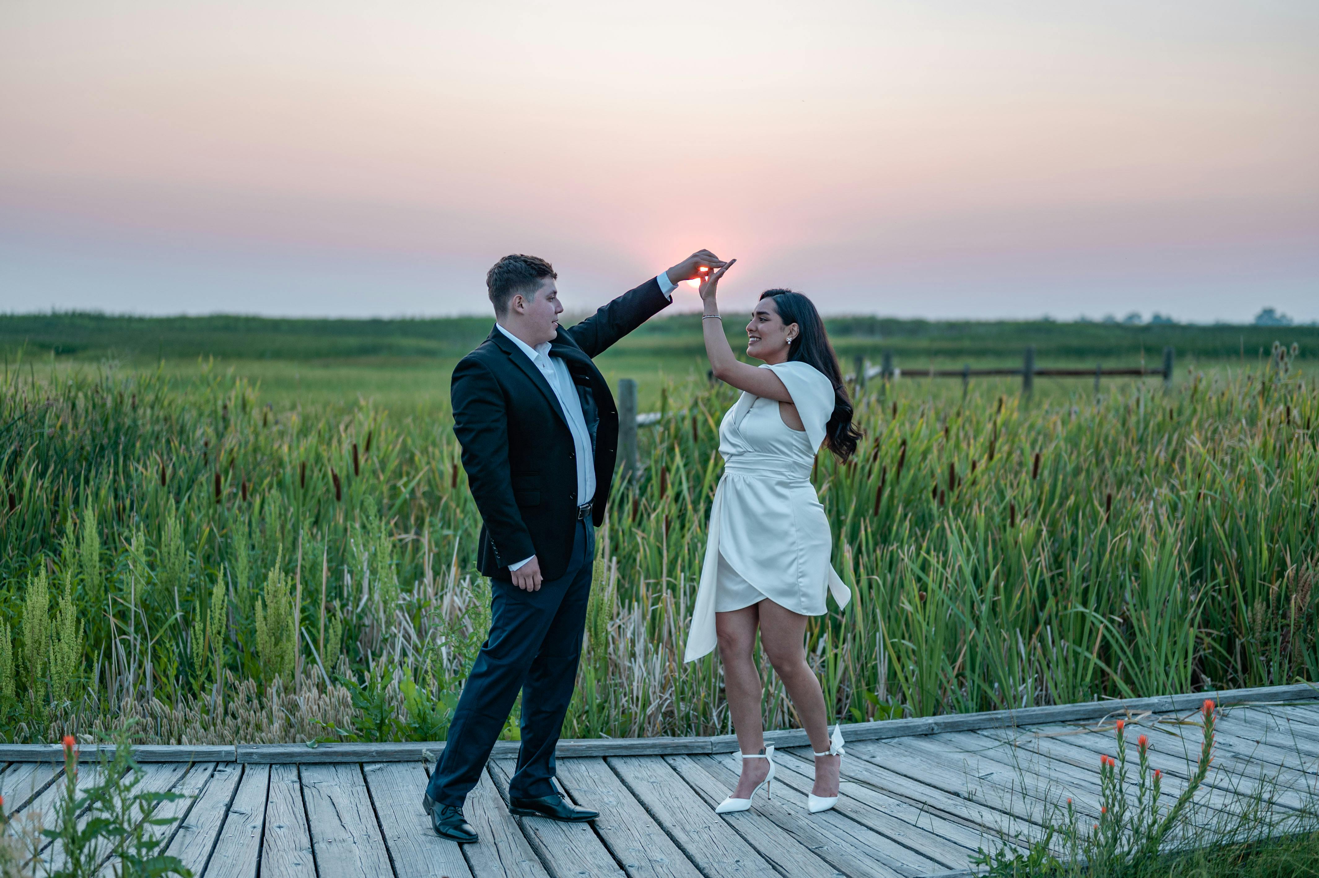 A couple elegantly dressed enjoys a joyful dance on a wooden bridge during sunset in nature.
