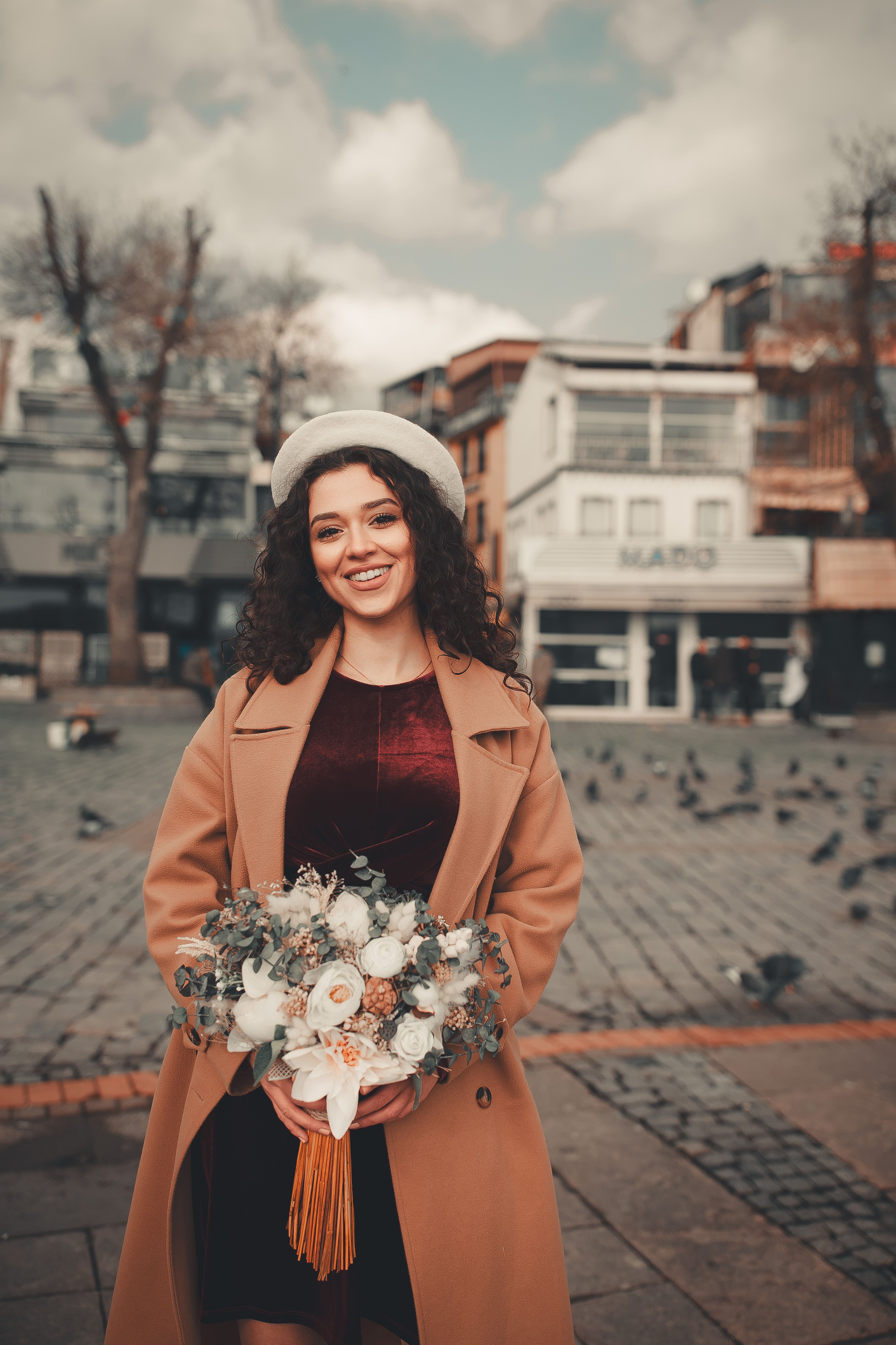 Smiling woman in a beige coat with a bouquet standing outdoors in a city square.