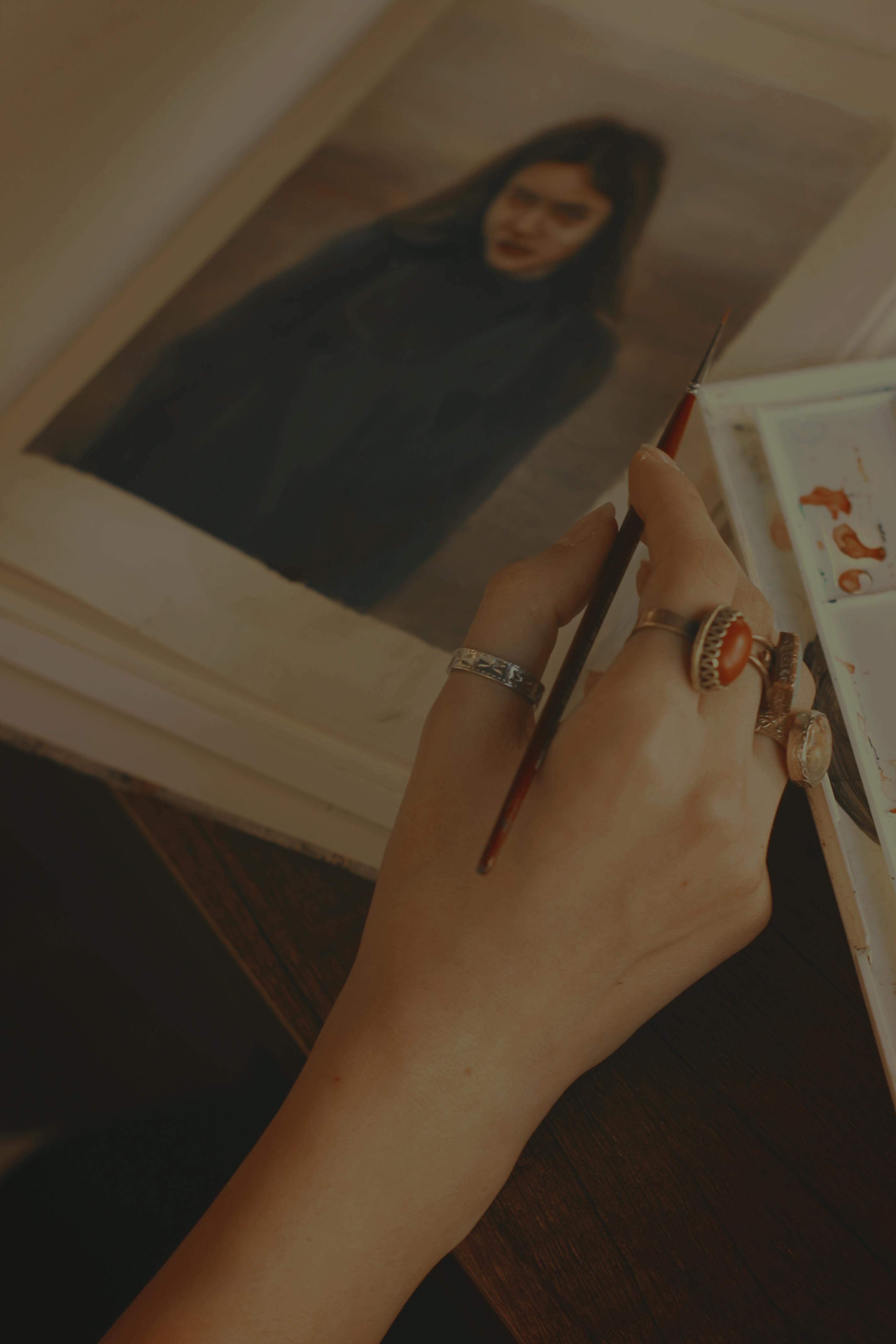 Close-up of a hand holding a paintbrush near a portrait painting, evoking a moody and artistic ambiance.