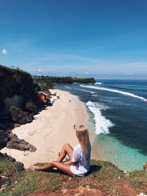 Free Woman enjoying the serene view from a cliff overlooking the pristine beach and ocean waves in Bali, Indonesia. Stock Photo