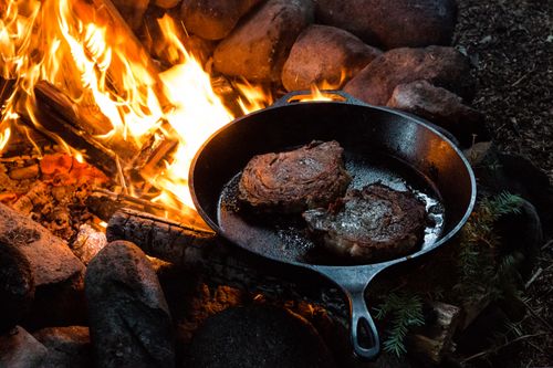 Free Sizzling steaks cooking in a cast iron pan over a campfire surrounded by logs and rocks. Stock Photo