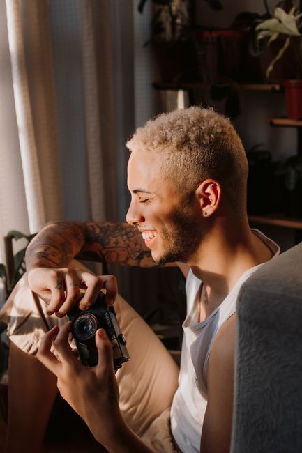 A smiling young man sits indoors, holding a camera, enjoying the warm sunlight.