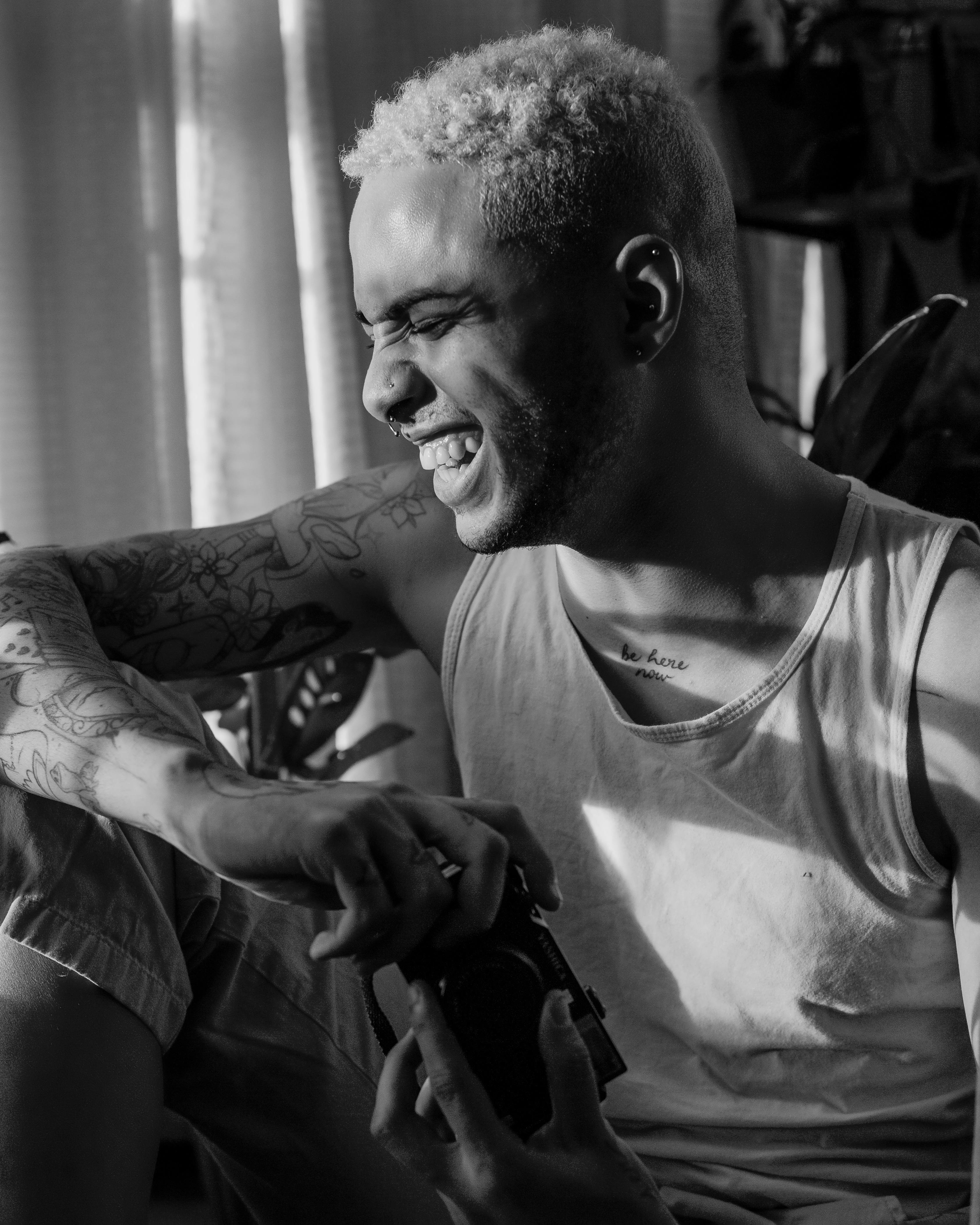 A smiling man with tattoos enjoys the sunlight indoors, holding a camera.