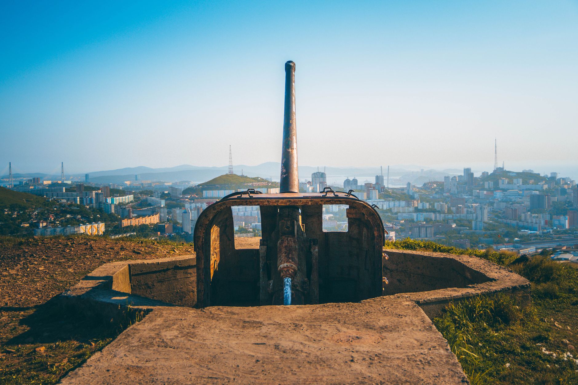 A rusted artillery installation with a city skyline in the background, captured outdoors during the day.