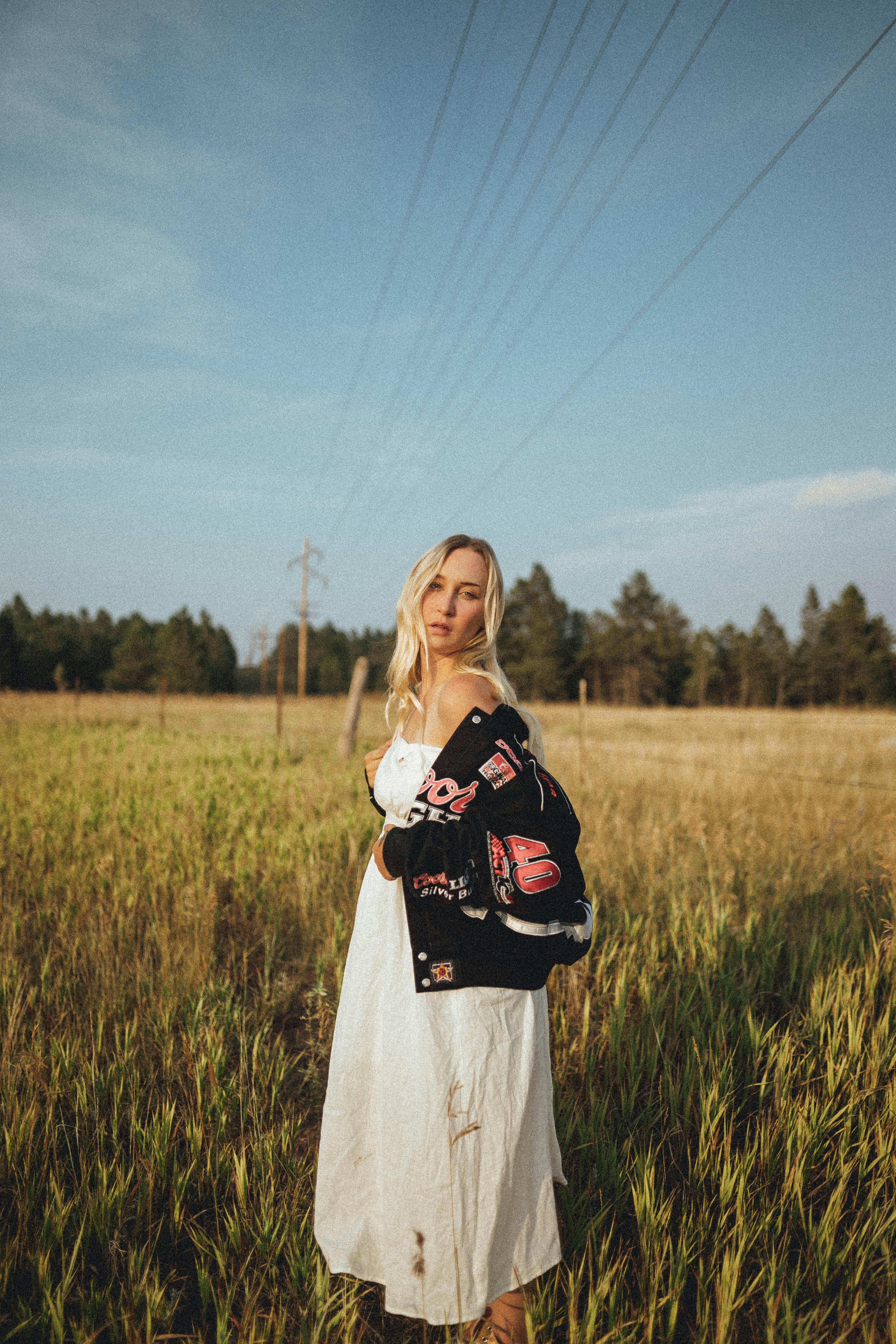 Woman posing in a field, wearing a black jacket and white dress under a blue sky.
