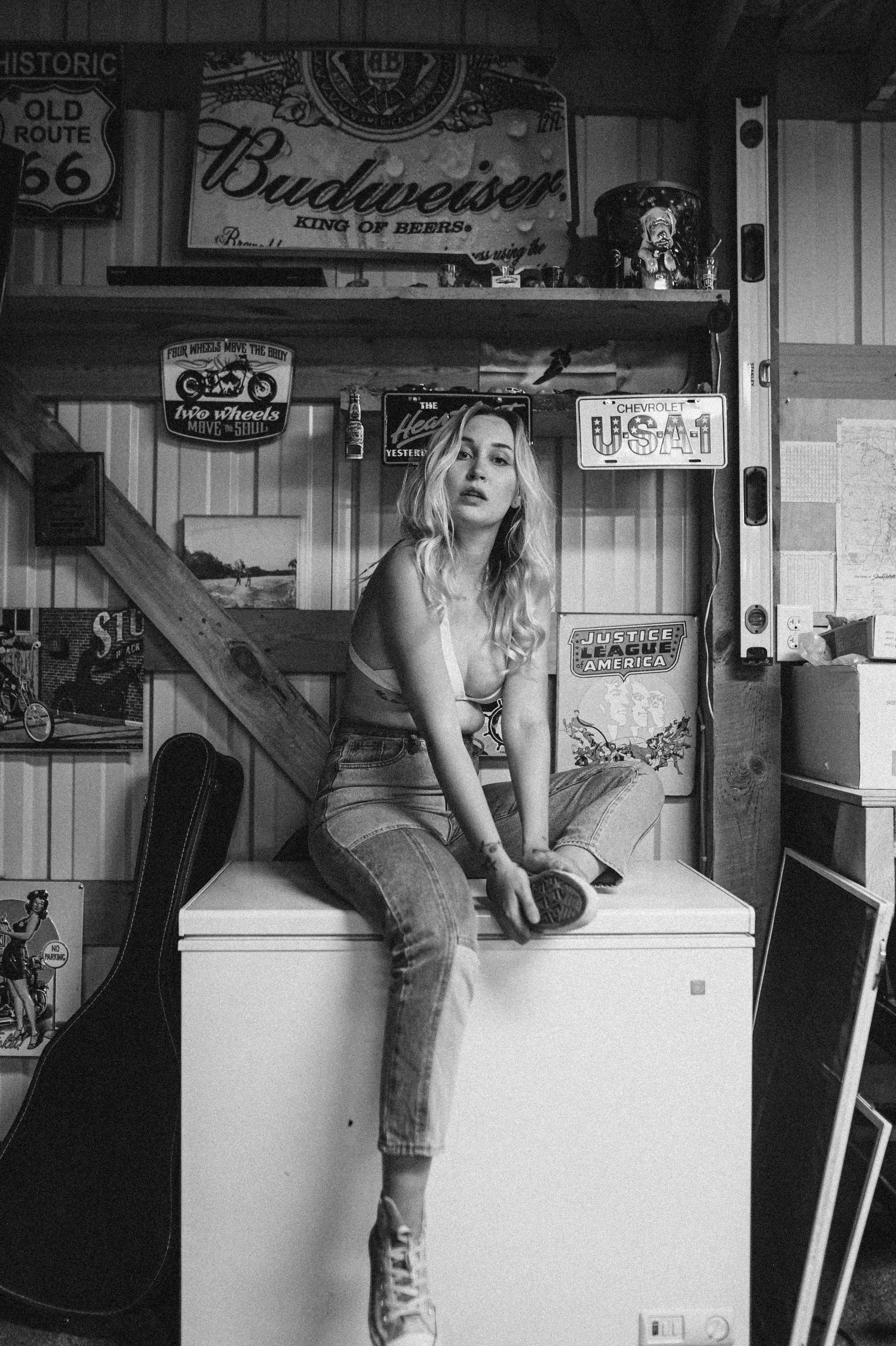 Black and white photo of a woman sitting on a freezer with vintage signs.