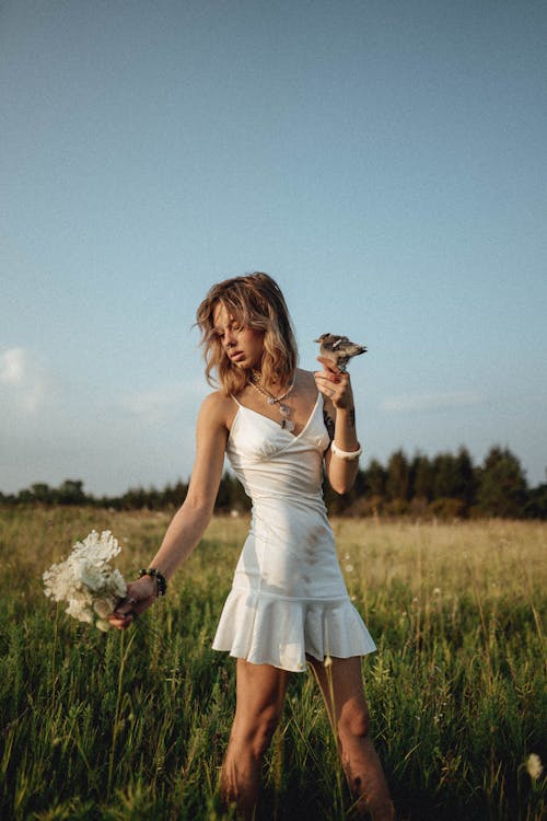 A Woman in White Dress Standing on the Grass Field while Holding Flowers