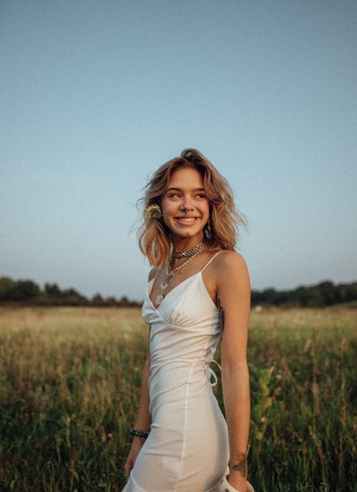 Woman in White Spaghetti Strap Dress Standing on Green Grass Field