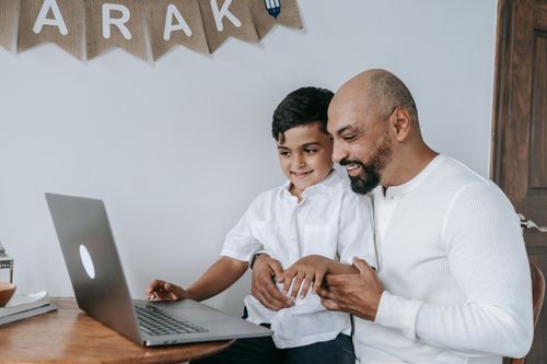 Free A father and son smiling while using a laptop, creating a moment of joy and bonding indoors. Stock Photo