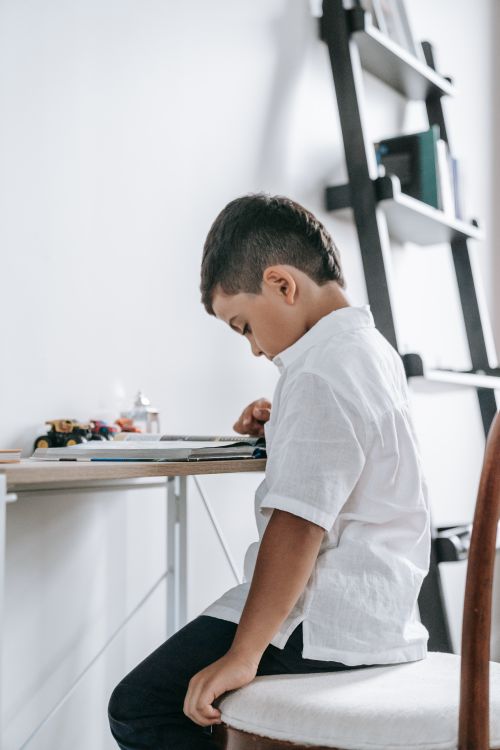 Free A young boy in a white shirt is focused on reading at a desk indoors, surrounded by toys and books. Stock Photo