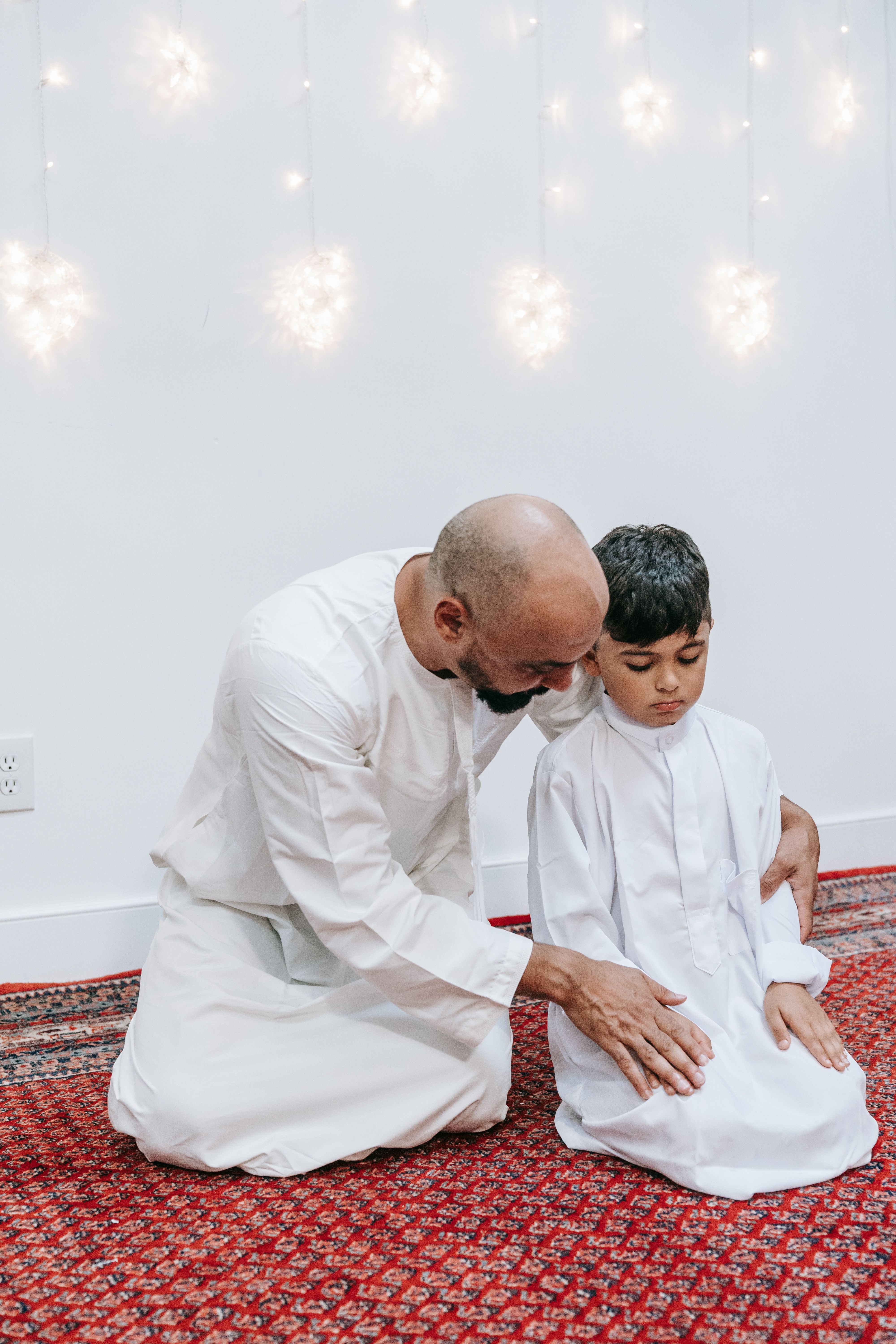 A father kneels with his son in prayer, both wearing white robes, against a backdrop of string lights.