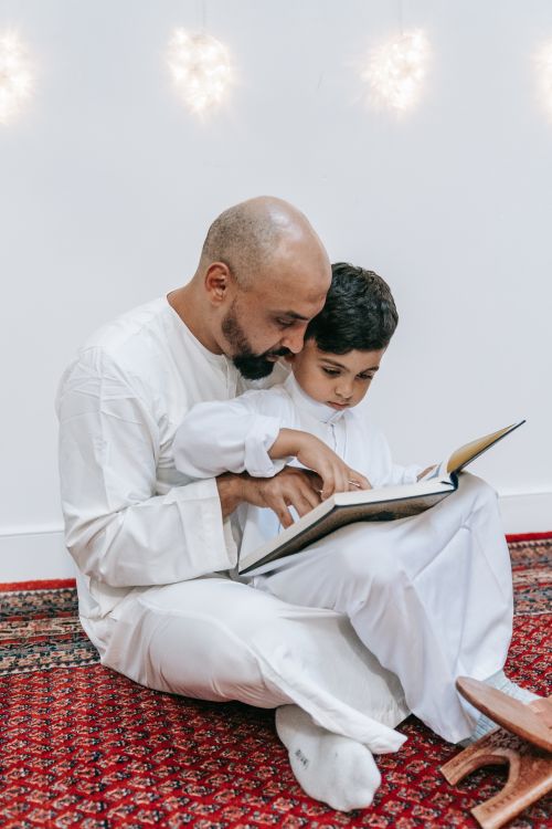 Free A father and son wearing traditional attire reading a book together inside a cozy room. Stock Photo