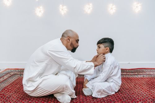 Free A father gently adjusting his son's collar, sitting on a prayer mat indoors. Stock Photo