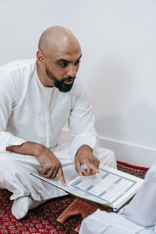 Free Man sitting indoors in a thobe, reading a religious text from Quran on a wooden stand. Stock Photo