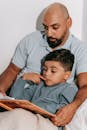 Man in Light Blue Shirt Reading a Book with a Boy in Gray Long Sleeve Shirt