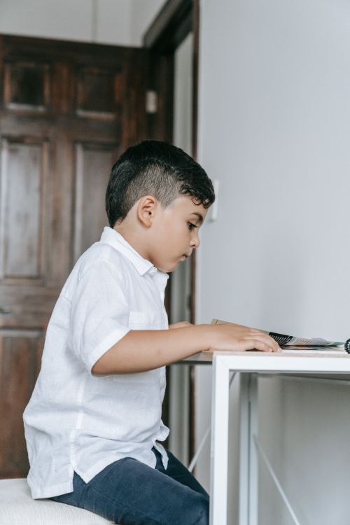 Free A young boy in a white shirt reading at a table indoors, focused and engaged. Stock Photo