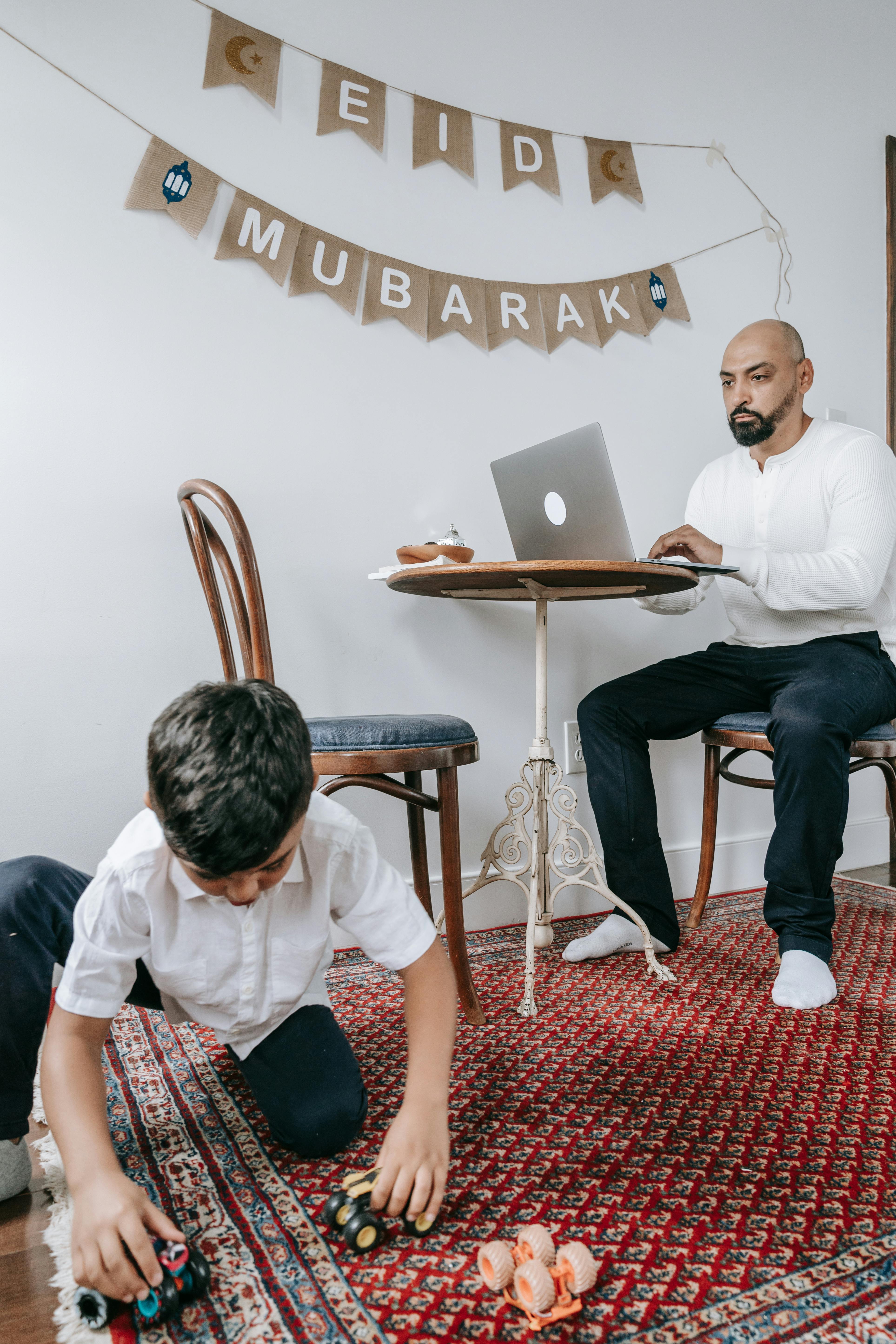 photo of a boy playing on the carpet and his father sitting with a laptop