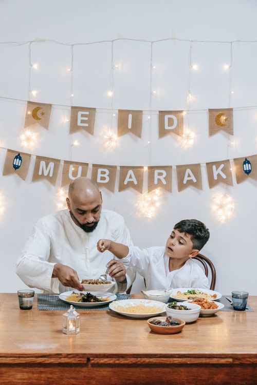 Free Father and son enjoying Eid dinner together indoors, decorated with festive lights. Stock Photo