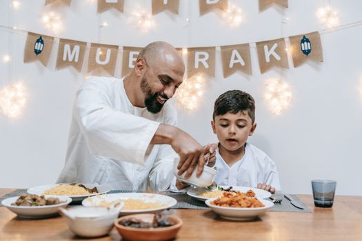 Father and son share a joyful Eid meal at a festively decorated table indoors.