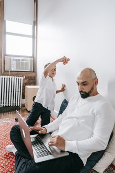 A father works on his laptop while spending quality time with his son in a cozy room.