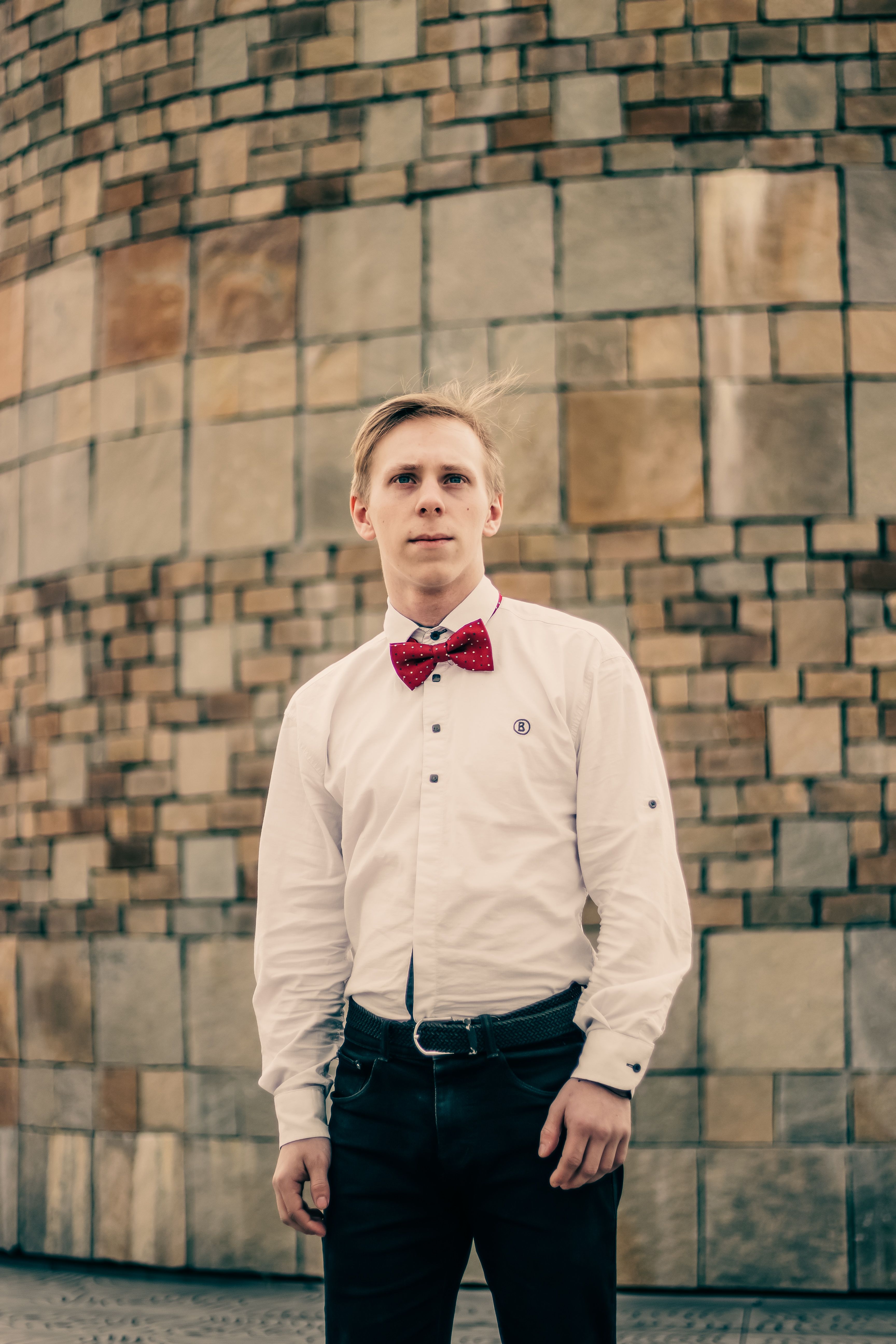 Young man in white shirt and red bowtie standing by a brick wall.