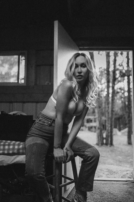 Black and white portrait of a woman in denim jeans sitting indoors on a wooden stool.