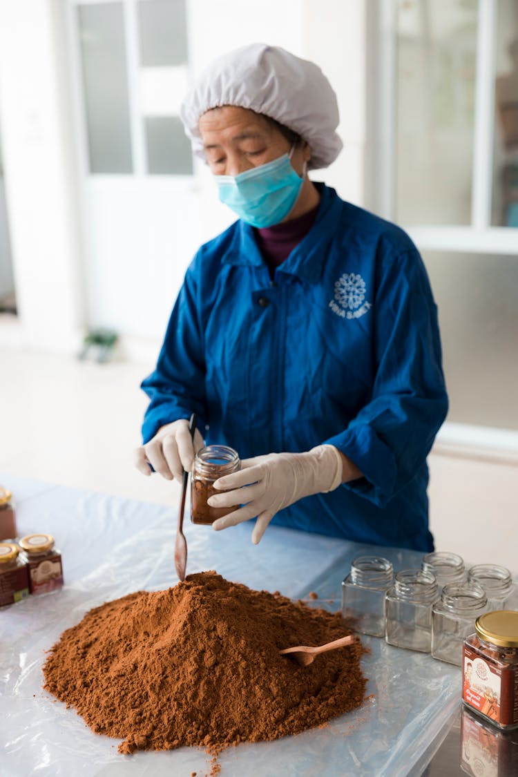 A Woman With Haircap And Mask Storing Brown Powder In Bottles