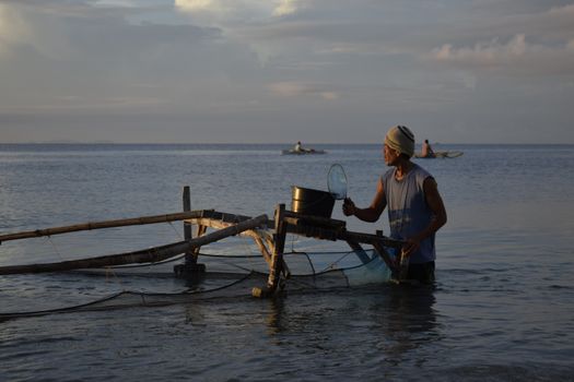 A fisherman stands with a net, fishing in calm coastal waters during a serene evening. - Fotografer Sidoarjo