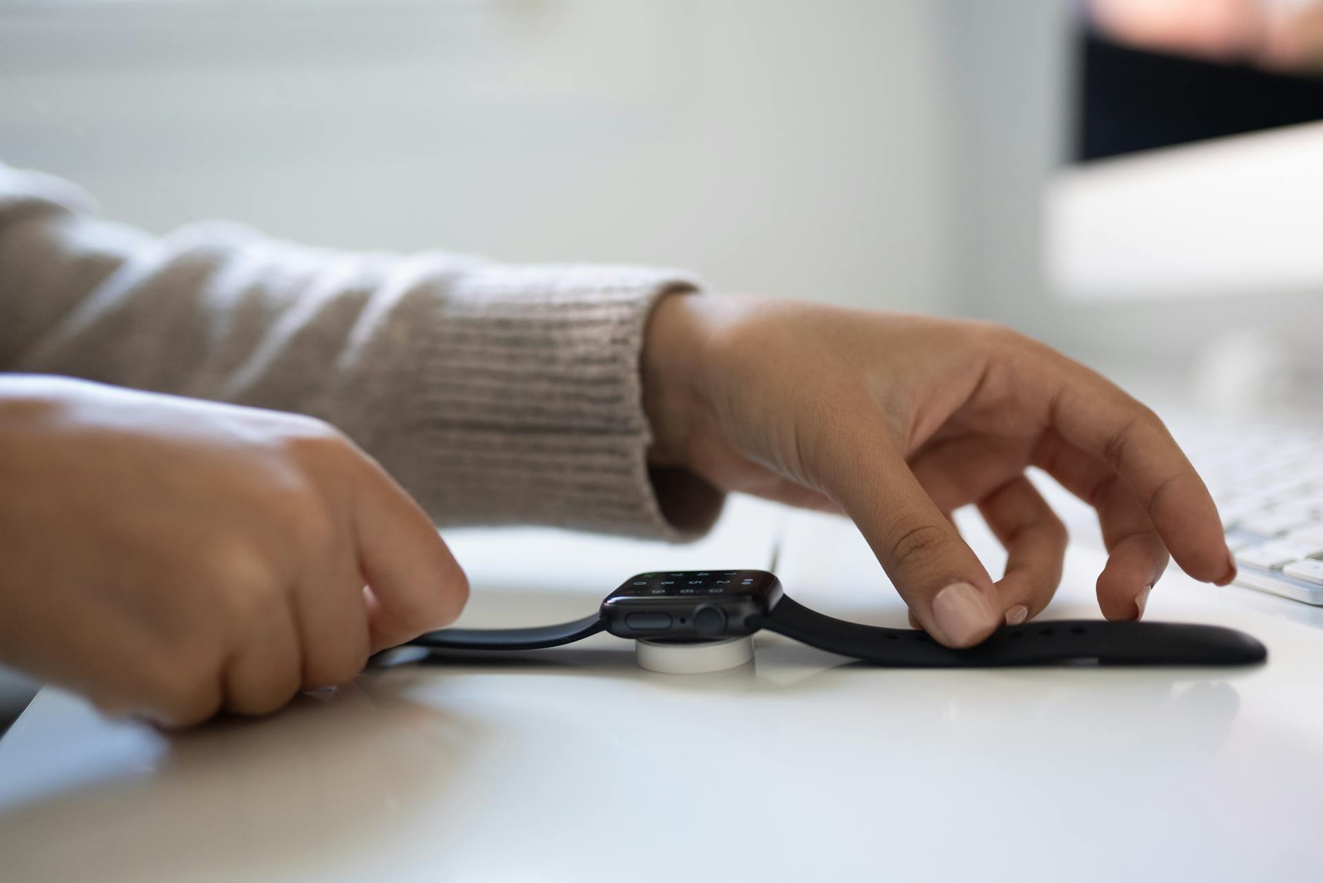 Close-up of hands placing a smartwatch on a charging dock on a white desk indoors.