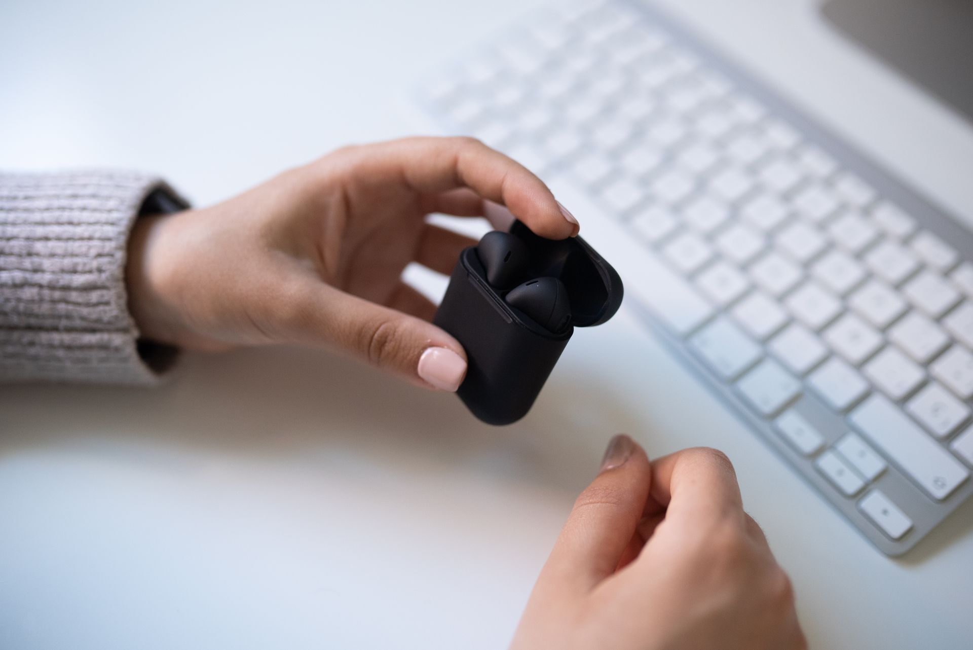 Close-up of hands holding black wireless earbuds near a computer keyboard, showcasing modern lifestyle and technology.